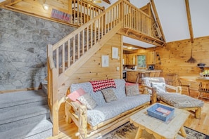 A wooden cabin interior with a stone wall, wooden staircase, sofa, chairs, and a dining table.