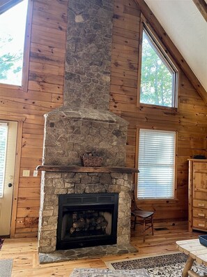 A log cabin interior with a stone fireplace, wooden walls, and large windows.