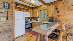 A rustic cabin interior with wooden walls, a wooden dining table, a refrigerator, a stove, and a window with a view of trees. - Cabin #1 - Fantasy Hideaway (Stanardsville)