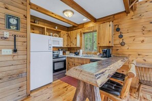 A rustic cabin interior with wooden walls, a wooden dining table, a refrigerator, a stove, and a window with a view of trees.