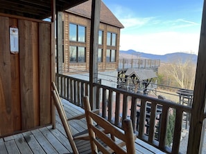 A wooden deck with a wooden chair, a wooden building with multiple windows, and a view of mountains in the background.