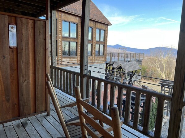 Une terrasse en bois avec une chaise en bois, un bâtiment en bois doté de nombreuses fenêtres, et une vue sur des montagnes en arrière-plan.