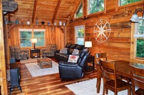 A wooden cabin interior with a sofa set, a dining table, and a large window.