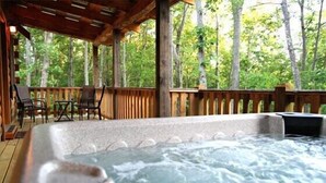 Een hottub op een houten terras met stoelen en een tafel, omringd door bomen.