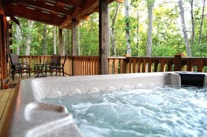 A hot tub on a wooden deck with chairs and a table, surrounded by trees.