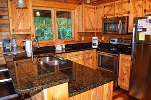 A kitchen with wooden cabinets, a granite countertop, a stainless steel refrigerator, and a window with a view of greenery.
