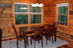 A wooden cabin interior with a dining table and chairs, large windows, and a chandelier.
