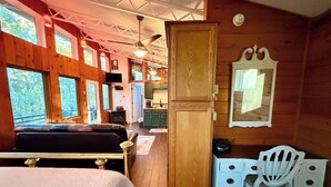 A cabin interior with a bed, a desk with a chair, a mirror, and a kitchen area visible through an open door. - Second Wind Mountain Cabin (Stanardsville)