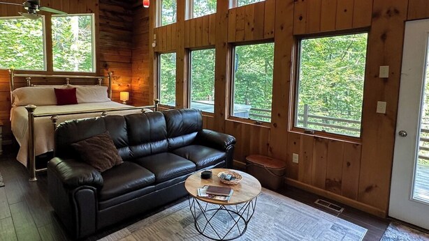 A log cabin interior with a bed, leather sofa, and large windows. - Second Wind Mountain Cabin (Stanardsville)
