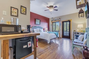 Quarto com cama, escrivaninha com micro-ondas, geladeira, ventilador de teto e janela com vista para árvores.