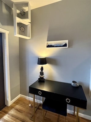 A corner room with a black desk, a lamp, a framed picture, and a white shelf with decorative items.