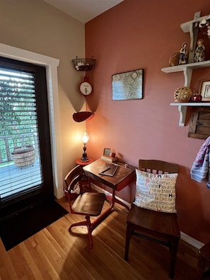 A room with a wooden floor, a desk with a lamp, a chair, a bookshelf, and a window with blinds.