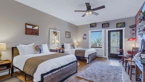 Une chambre avec deux lits, un ventilateur de plafond, un bureau avec une chaise et une porte donnant sur l’extérieur.