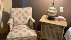 A floral-patterned armchair next to a wooden nightstand with a lamp and a clock. - First Lady - Rm. 206 (Stanardsville)