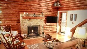 Ein Blockhaushaus-Interieur mit Kamin aus Stein, Holzwänden, einer Couch, einem Sessel und einem Fernseher.