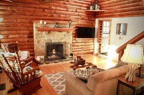 Interior de una cabaña de troncos con chimenea de piedra, paredes de madera, un sofá, un sillón y un televisor.