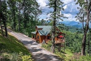 A wooden cabin with a porch, surrounded by trees and a gravel path.