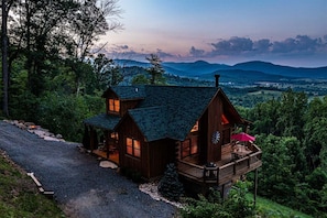 A wooden cabin with a deck, surrounded by trees and mountains.