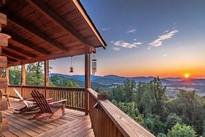 A wooden deck with rocking chairs, a covered area, and a view of the sunset over a mountainous landscape.