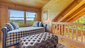 Una cabaña de madera con habitación en entrepiso, que incluye una cama, un banco acolchado y un ventanal con vistas al paisaje.