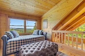 A wooden cabin with a loft bedroom featuring a bed, a cushioned bench, and a large window offering a view of the landscape.