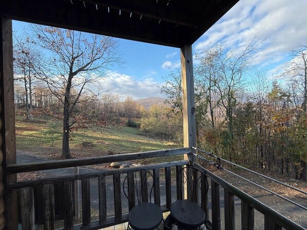 A covered outdoor area with a wooden railing, two black chairs, and a view of a forested landscape.