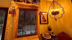 Une pièce avec une fenêtre en bois munie de stores, une étagère décorative sur laquelle sont disposés divers objets, un luminaire fixé au mur et un tableau encadré.