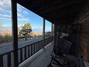 A wooden porch with a railing, a rocking chair, and a view of a road and trees.