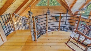 A wooden interior with a curved staircase, a rocking chair, and a view of greenery outside. - Cabin #10 - Our Mountain Cabin (Stanardsville)
