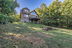A two-story log cabin with a stone foundation, a covered porch, and a grassy yard with scattered leaves.
