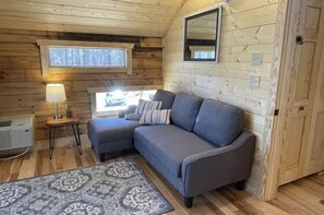 A wooden cabin interior with a gray sofa, a patterned rug, a window with a view, and a wall-mounted mirror. - Speckled Owl Mountain Cabin (Stanardsville)