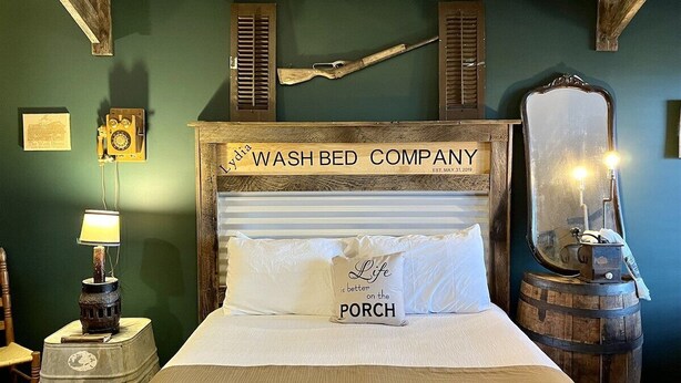 En seng med et trægavl med teksten 'WASH BED COMPANY', en pude med teksten 'Life is better on the PORCH', en vintage lampe og et spejl på en træfad.