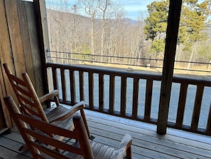 A wooden deck with a wooden rocking chair and a railing overlooking a forested area.