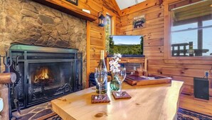 L’intérieur d’un chalet en bois est doté d’une cheminée en pierre, d’un téléviseur qui diffuse des images de paysages et d’une table en bois sur laquelle sont posés des verres à vin.