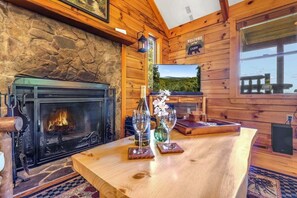 Interior de una cabaña de madera con chimenea de piedra, un televisor mostrando una vista panorámica y una mesa de madera con copas de vino.