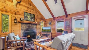 L’intérieur d’une cabane en rondins, avec une cheminée en pierre, des meubles en bois et une vue sur l’extérieur par une porte.