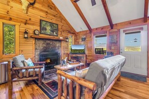 A log cabin interior with a stone fireplace, wooden furniture, and a view of the outdoors through a door. - Mountain View Hideaway (Stanardsville)