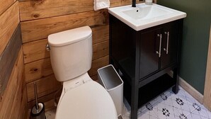 Ein Badezimmer mit Holzvertäfelung, einer weißen Toilette, einem schwarzen Waschbecken mit Spiegel darüber und einem Mülleimer.
