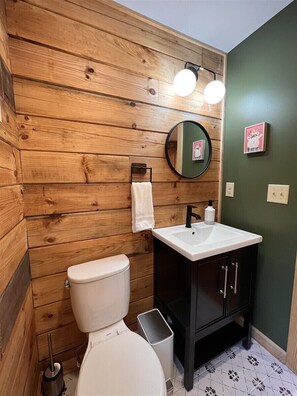Une salle de bain avec un lambris en bois, des toilettes blanches, un lavabo noir avec un miroir au-dessus, et une poubelle.