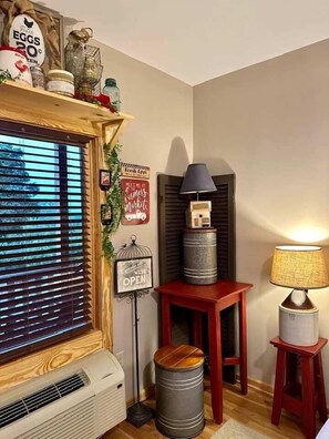 A corner room with a window, a radiator, a lamp, a small table, and decorative items. - Farmer`s Market Rm. 102 (Stanardsville)