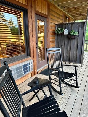 A wooden cabin with a porch, two rocking chairs, a table, and a window with blinds. - Farmer`s Market Rm. 102 (Stanardsville)
