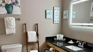 A bathroom with a toilet, a wooden towel rack, a sink with a mirror, and a framed floral artwork on the wall. - Once Upon A Time Rm. 103 (Stanardsville)