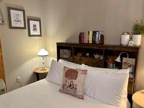 Ein ordentlich bezogenes Bett mit einem Zierkissen, einer Nachttischlampe, einem Bücherregal mit Büchern und gerahmten Bildern an der Wand.