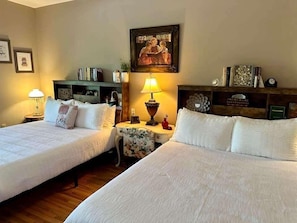 A hotel room with two beds, a small table with a lamp, a bookshelf, and a framed picture on the wall.