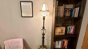 A corner room with a wooden floor, a brown chair with a blanket, a tall floor lamp, and a bookshelf filled with books. - Once Upon A Time Rm. 103 (Stanardsville)