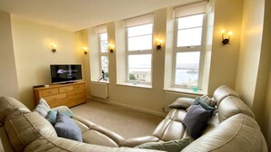 A living room with a beige leather sofa, a wooden TV stand, and a large window with a view of a body of water and buildings. - Two Bed Apartment with a Millionaire's sea view (Torquay)
