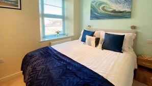 A bedroom with a bed, a window with blinds, a painting of a wave, and a nightstand with a lamp. - Two Bed Apartment with a Millionaire's sea view (Torquay)