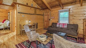 L’intérieur d’un chalet en bois, avec un canapé, des fauteuils, une petite table et une fenêtre équipée de stores.