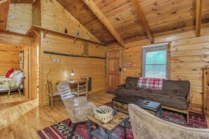 Interior de una cabaña de madera con un sofá, sillas, una mesita y una ventana con persianas.