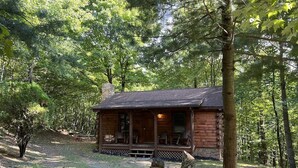 Une cabane en rondins dotée d’un porche, entourée d’arbres et bordée d’un chemin de gravier.